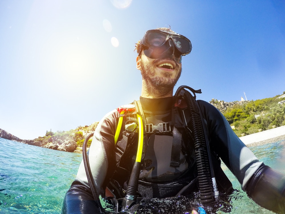 Smiling diver wearing goggles and scuba equipment waist deep in clear water near a shoreline