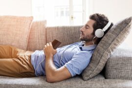 man reclined on sofa wearing headphones with eyes closed holding music player